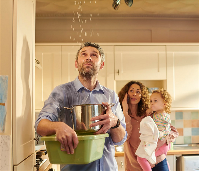 a man holding a bucket to catch water falling from the ceiling while his wife and child are in the background on the phone